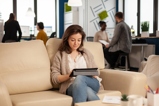 Employee taking break sitting on confortable couch holding tablet browsing, reading great news and smiling. Multiethnic creative coworkers planning new financial project working in open plan office