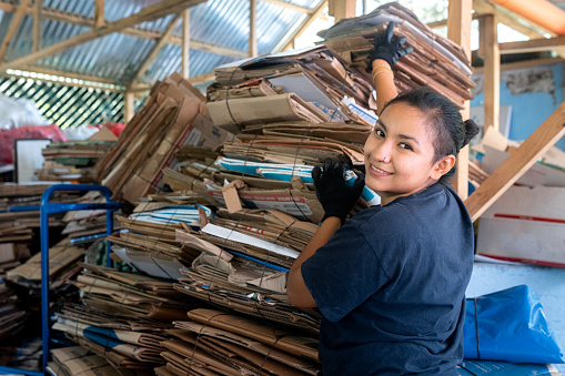Hispanic woman stacks cardboard, glances at camera, showcasing focus and dedication at recycling site