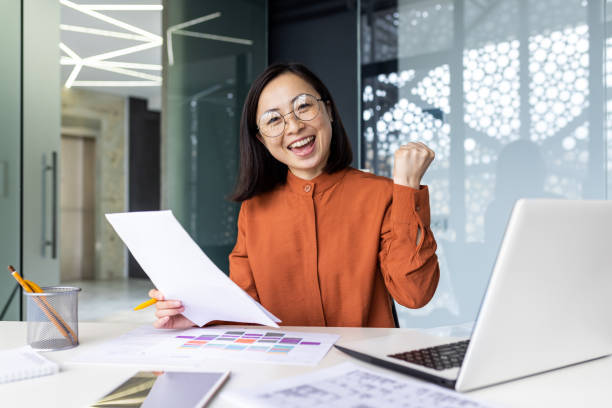 istock Happy and successful satisfied woman at workplace working with documents and papers, business woman smiling and looking at camera, portrait of asian woman got successful achievement results 1864183048