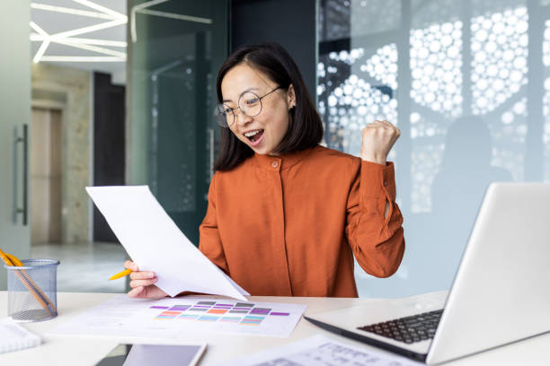 istock Happy and successful satisfied woman at workplace working with documents and papers, asian business woman received successful achievement results, holding hand up celebrating victory and triumph 1864183049