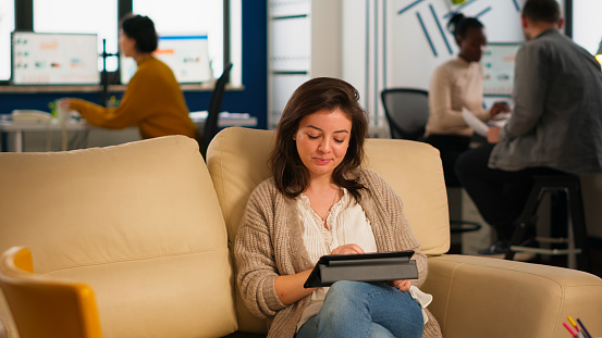 Employee taking break sitting on confortable couch holding tablet browsing, reading great news and smiling. Multiethnic creative coworkers planning new financial project working in open plan office