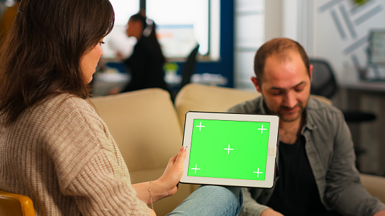 Business people sitting on couch analysing financial statistics, holding tablet with green screen while diverse team working on background. Multiethnic coworkers planning project on chroma key display