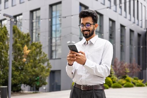 Young successful businessman walking down the street holding a phone in his hands, an Indian man using an application on a smartphone smiling contentedly from outside an office building.