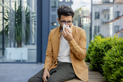 A sick man sneezes sitting on a bench outside an office building in the air, a cold businessman with the flu has a runny nose.