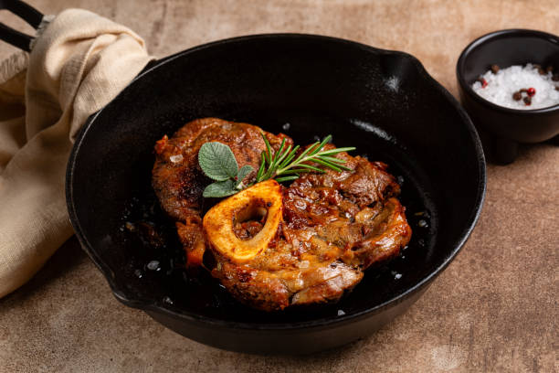 Braised beef steak ossobuco, meat on the bone, cooked with onion, salt, spices, rosemary and sage in an iron cast pan and brown concrete background. Braised beef steak ossobuco, meat on the bone, cooked with onion, salt, spices, rosemary and sage in an iron cast pan and brown concrete background. ossobuco stock pictures, royalty-free photos & images