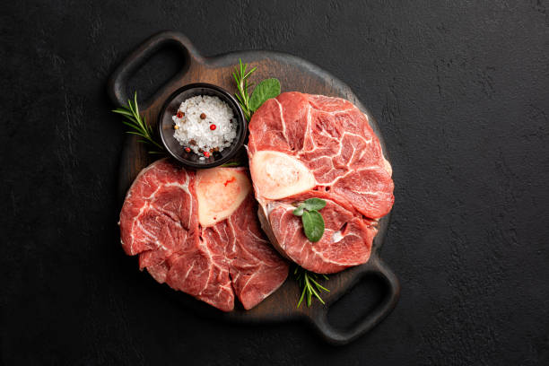 Top view of Raw beef steak ossobuco, meat on the bone, with salt, spices, rosemary and sage on a cutting board and black textured concrete background. Top view of Raw beef steak ossobuco, meat on the bone, with salt, spices, rosemary and sage on a cutting board and black textured concrete background. ossobuco stock pictures, royalty-free photos & images