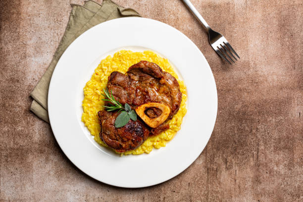 vista superior do prato com prato italiano ossobuco, carne refogada servida com risoto de açafrão alla milanese, especialidade da cozinha lombarda. fundo marrom. - veal meat osso buco beef stew - fotografias e filmes do acervo