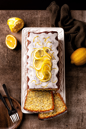 Sliced pound cake with lemon zest, sugar and lemon glaze and poppy seeds on a white plate and brown background. Dark mood.
