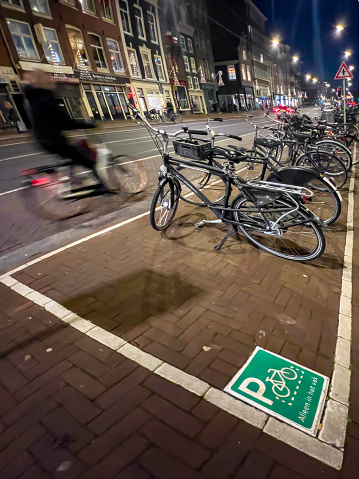 Blurred movement of a cyclist and many parked bicycles. Looking down at a green and white sign indicating bicycle parking on a cobbled street in Amsterdam with text in Dutch 