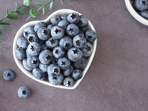 Organic ripe fresh blueberries in heart shape white bowl on grey background. Concept for healthy eating and nutrition, Top view with space for your text. Superfood.
