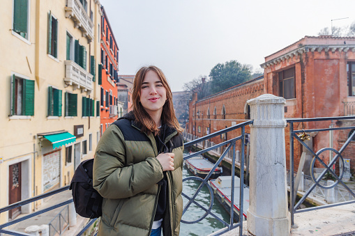 Portrait of beautiful teenage girl in winter jacket standing on a pedestrian bridge over a canal and smiling at camera, visiting Venice, Italy