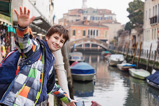 Preadolescent boy in winter jacket standing by canal with moored boats and waving at camera, traveling to Venice, Italy