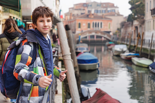 Preadolescent boy in winter jacket standing by canal with moored boats and showing thumbs up to camera, traveling to Venice, Italy
