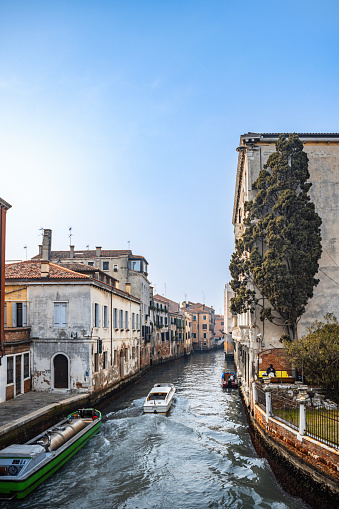Secondary canal between pier sidewalks and residential buildings rising from the water with boat driving by in Venice, Italy