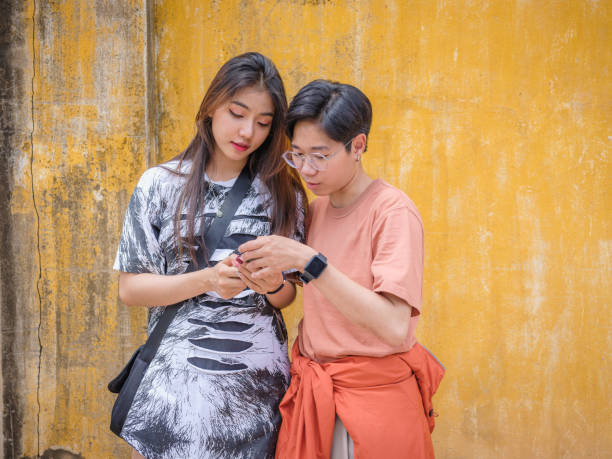 Young Asian friends using mobile phone and standing at yellow old wall in background, Happy friends relaxing and spending time together at outdoors, People and lifestyle