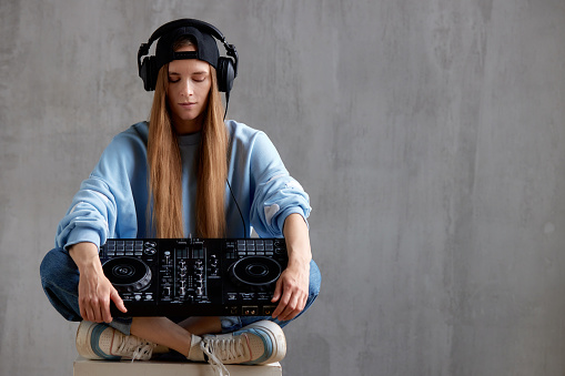 Studio portrait of a young pretty long-haired DJ girl in a blue sweater, loose jeans and a black baseball cap posing while sitting with a DJ console in her hands. Music, fun and creativity.