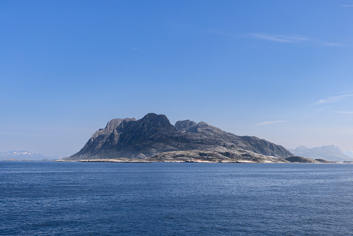 A serene summer tableau captured from a ferry traveling to Lofoten Island, illustrating a solitary, rugged mountain rising above the placid North Sea, framed by a radiant, sapphire sky in Norway