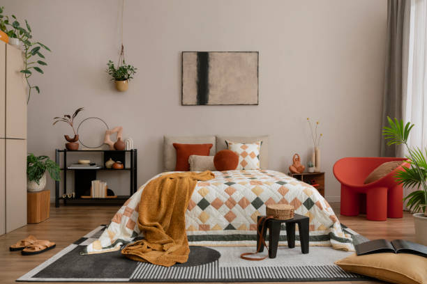istock Warm and cozy bedroom interior with mock up poster frame, beige and orange bedding, patterned rug, black rack, plants, red armchair, plaid, curtain and personal accessories. Home decor. Template. 1864330799