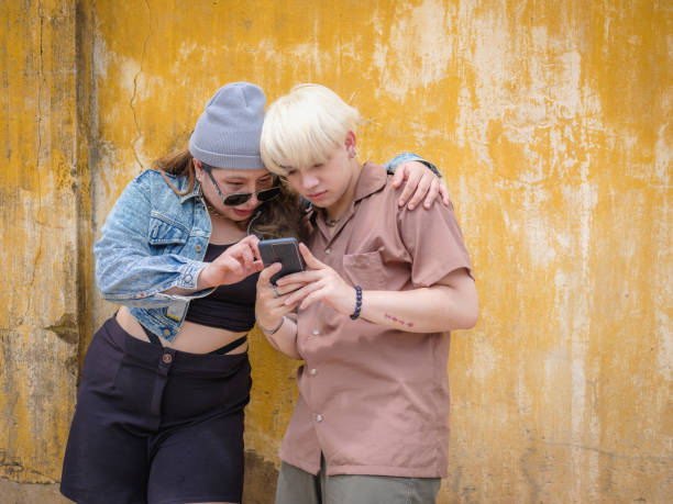 Young Asian friends using mobile phone and standing at yellow old wall in background, Happy friends relaxing and spending time together at outdoors, People and lifestyle
