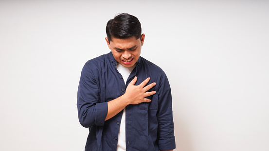 Young Asian man touching his chest with pain expression. Heart attack gesture on white background