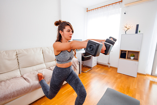 Active young woman in sportswear doing lunges using weights while working out at home