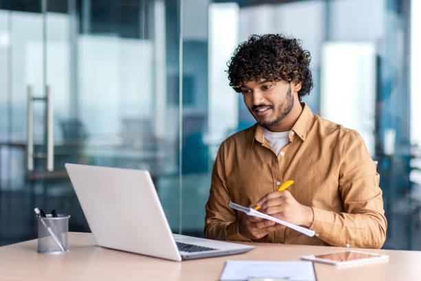 istock Man studying online remotely, businessman improving qualifications at the workplace inside office, writing down data in a notebook, watching a video course, satisfied with high results, smiling 1864417490
