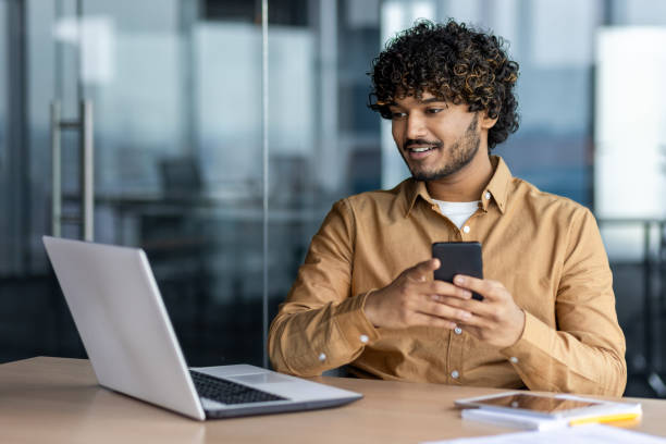 istock Young successful hispanic businessman using app on phone, man smiling contentedly inside office at workplace, employee working with laptop 1864418056