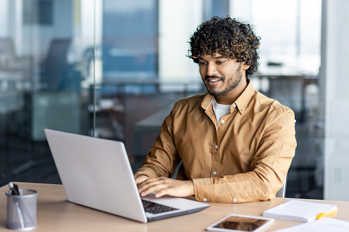Happy and smiling hispanic businessman typing on laptop, office worker with curly hair happy with achievement results, at work inside office building