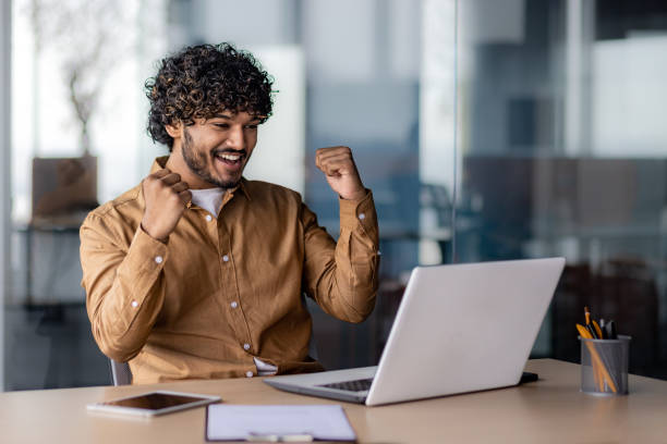 istock Successful hispanic businessman programmer celebrating victory and triumph, man inside office sitting at table and working using laptop, holding hands up gesture of achieving good results 1864449625