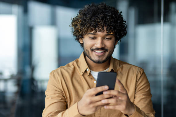 istock Close up of young businessman using online application on phone, man inside office reading good news from smartphone, latin american man smiling contentedly 1864449629