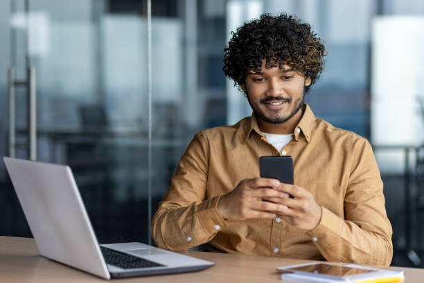 istock Successful young businessman inside office at workplace, man typing message and browsing social media, hispanic smiling sitting holding phone, browsing online sites 1864450157