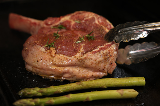 A tomahawk or ribeye steak on the bone, just placed on the griller or bbq (barbecue, barbeque) with a serve of green asparagus. Metal tongs turning or placing the meat.