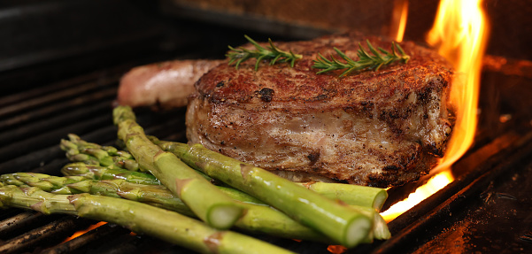 The prefect bone-in rib-eye steak cooking on the bbq, barbecue, barbeque or griller with flames and asparagus sides. Rosemary garnish and close-up angle.