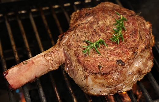 A single rib-eye steak on the bone with rosemary herb garnish cooking on the bbq or barbecue grill. The prefect cut of beef meat close up cooking on the griller.