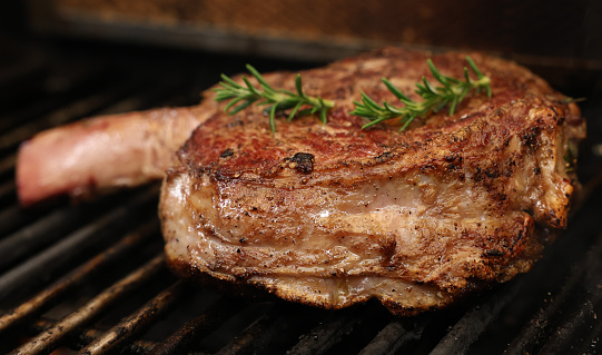 A single ribeye steak on the bone with rosemary herb garnish cooking on the bbq or barbecue grill. The prefect cut of beef meat close up cooking on the griller.