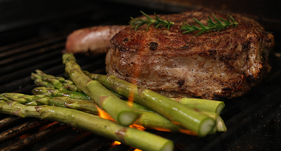 The prefect bone-in rib-eye steak cooking on the bbq, barbecue, barbeque or griller with flames and asparagis sides. Rosemary garnish and close-up angle.