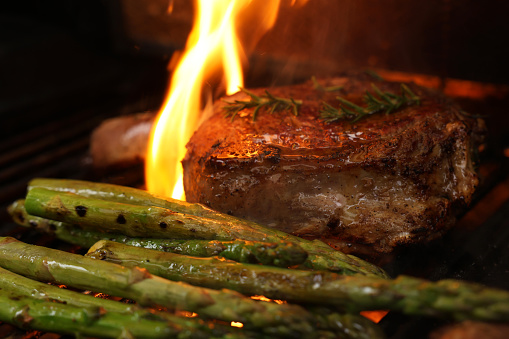 A mouth watering prefect bone-in rib-eye steak cooking on the bbq, barbecue, barbeque or griller with flames and asparagus sides. Rosemary garnish and close-up angle.