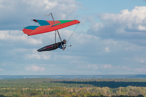 Colorful red hang glider wing soars over fields and forests with blue sky and clouds on the background.