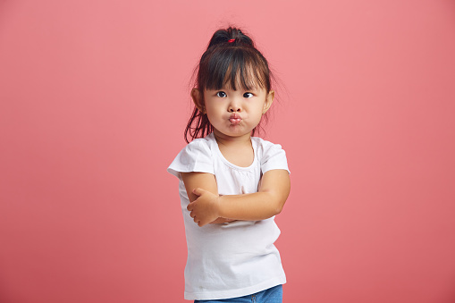 Offended little asian girl with insulted expression, scowls face, stick out lips in discontent, has a negative mood, dressed in white t shirt,crossed hands in front of her poses over pink isolated background.