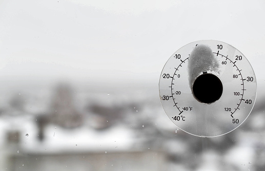 Window outdoor thermometer on glass in cloudy weather and snow cap. Concept of winter weather and cold snap, snow storm.