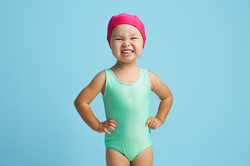 Cheerful and cute small asian girl in swim costume and cap prepares for floating in pool, dressed in swimwear, has healthy body, poses indoor against blue wall. Active rest for children.