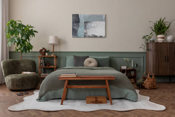 Creative composition of bedroom interior with mock up poster frame, green bedding, wooden side table, stylish bench, beige rug, plants, basket, lamp and personal accessories. Home decor. Template.