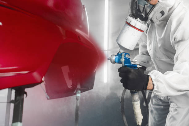 istock Car paint worker spraying red paint to car body element using spray gun in vehicle workshop chamber. Complex restoration of automobile body. 1864548616