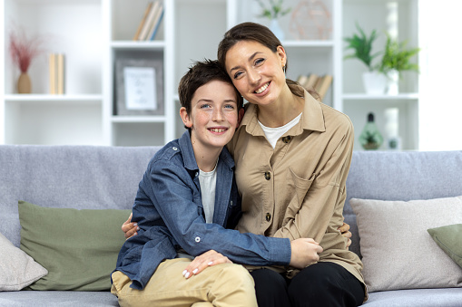 Portrait of family, mother and teenage son sitting on sofa at home and hugging, smiling at camera.