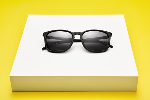 Black folded sun glasses is on white pedestal in showcase. Full rim stylish sunglasses is on flat box isolated on yellow background. Summer shopping, sale, vacation, touristic sunnies ads template.