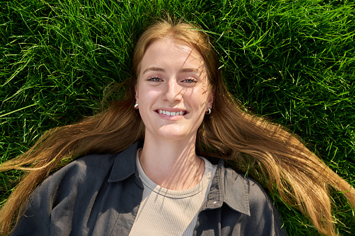Top view, close-up of face of smiling blonde teenage girl lying on grass. Beautiful positive female with long hair looking at camera. Adolescence, youth, beauty, lifestyle concept