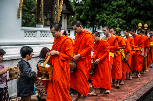 Luang Prabang, Laos - July 18, 2015: Luang Prabang, Laos - July 18, 2015: Morning alms in Luang Prabang, known as Tak Bat, is a serene Buddhist ritual. At sunrise, barefoot monks, draped in saffron robes, walk in silence through the streets. 

This photo encapsulates the moment as visitors graciously offer food and beverages to the monks.
