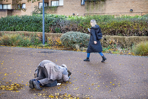 South Bank, London, England - November 13th 2023:  Beggar laying in a pitiful position on the ground and asking for money while people are walking by