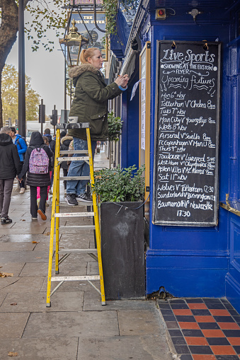 Euston Road, London, England - November 9th 2023:  Woman on a ladder setting up Christmas decoration outside a pub with a menu written on a blackboard
