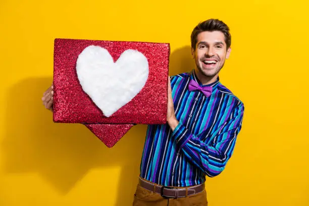 Photo of Photo of positive good mood man dressed striped shirt bow tie holding big social media heart box isolated on yellow color background